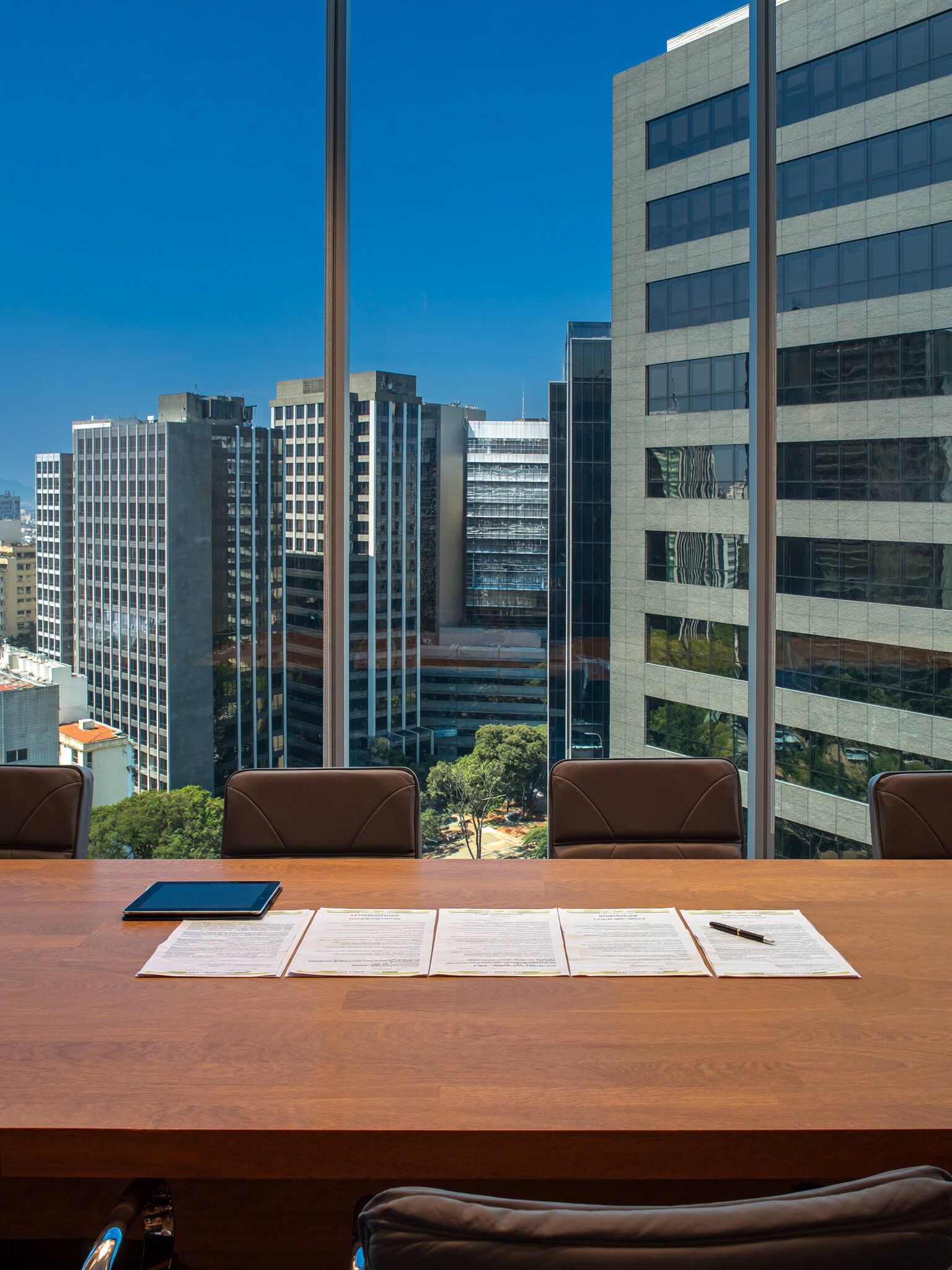 Sala de reunião iluminada pelo sol, com documentos sobre a mesa e prédios da cidade pela janela