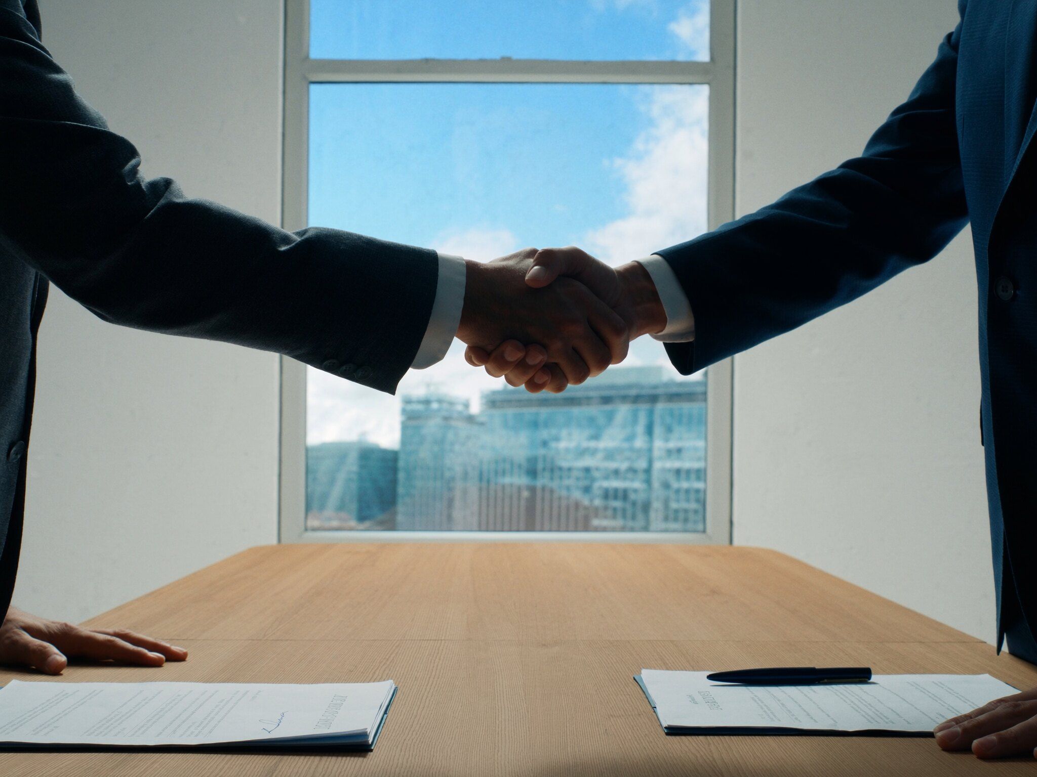 Aperto de mão sobre uma mesa de reunião com contratos assinados e uma caneta, em um escritório claro