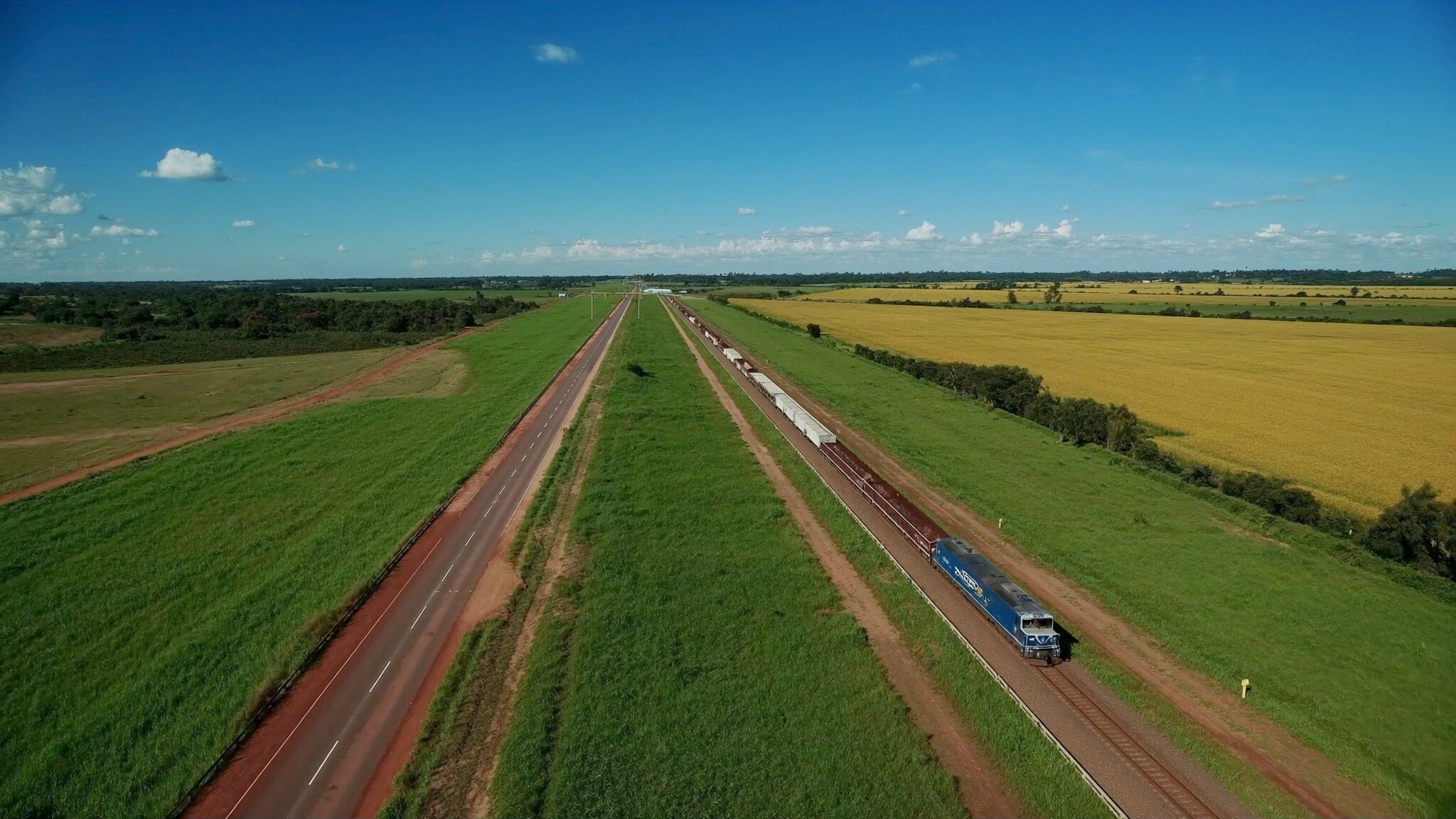 Rodovia e ferrovia lado a lado cruzando o interior do Brasil, com um trem de carga, em um dia de sol