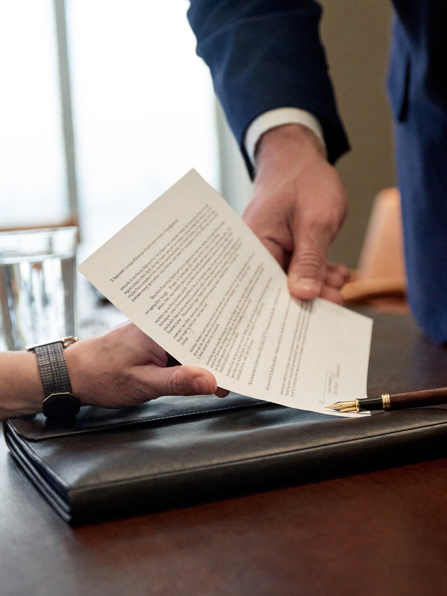 Mãos revisando um contrato impresso sobre uma mesa de madeira, com caneta-tinteiro e pasta de couro
